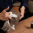Person cleaning a leather shoe with a white cloth on a wooden surface.