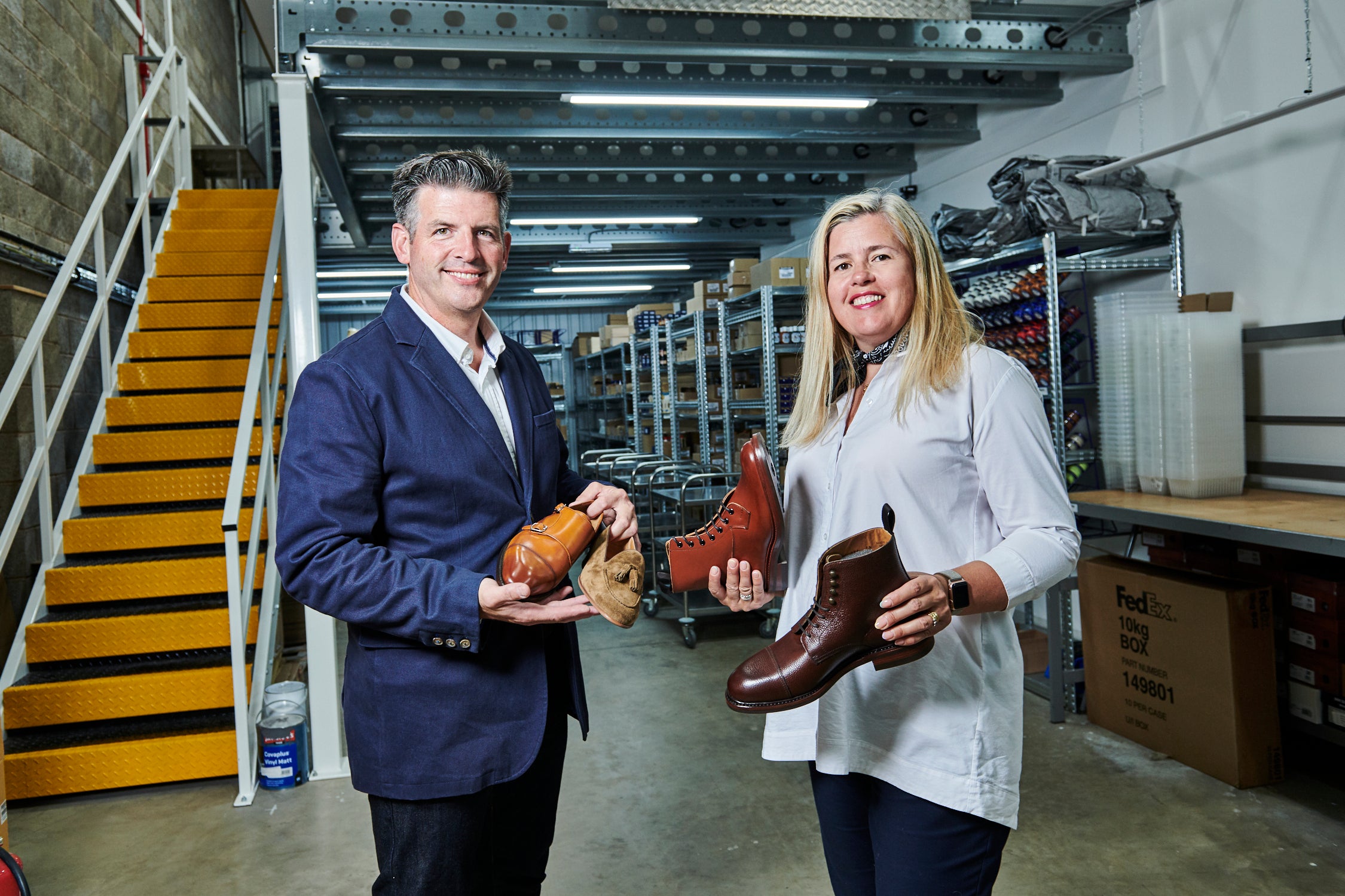 Julian & Sarah Nelson, Owners and Founders of A Fine Pair Of Shoes Ltd proudly stand in their modern, solar powered warehouse purpose built for the business smartly dressed holding Berwick 1707 & Trickers shoes and boots