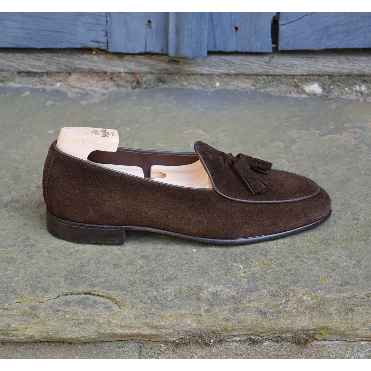 A dark brown suede tassel loafer with a Vegano Moka leather trim, displaying an almond-shaped toe and a fine ridged rubber sole.