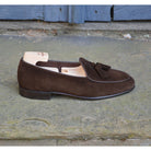 A dark brown suede tassel loafer with a Vegano Moka leather trim, displaying an almond-shaped toe and a fine ridged rubber sole.