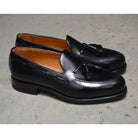 A black tasselled slip-on shoe with a polished, smooth upper and a classic round toe. The shoe is set against a natural stone steps background.