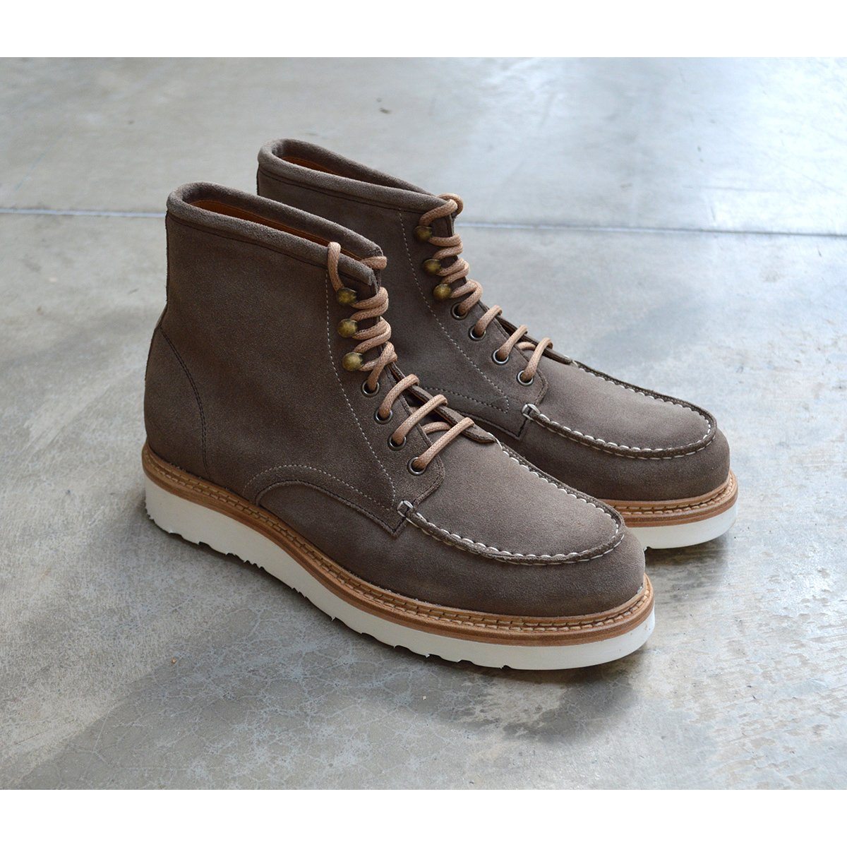A grey and tan suede apron boot with lace-up closure, featuring a white sole and a natural welt.