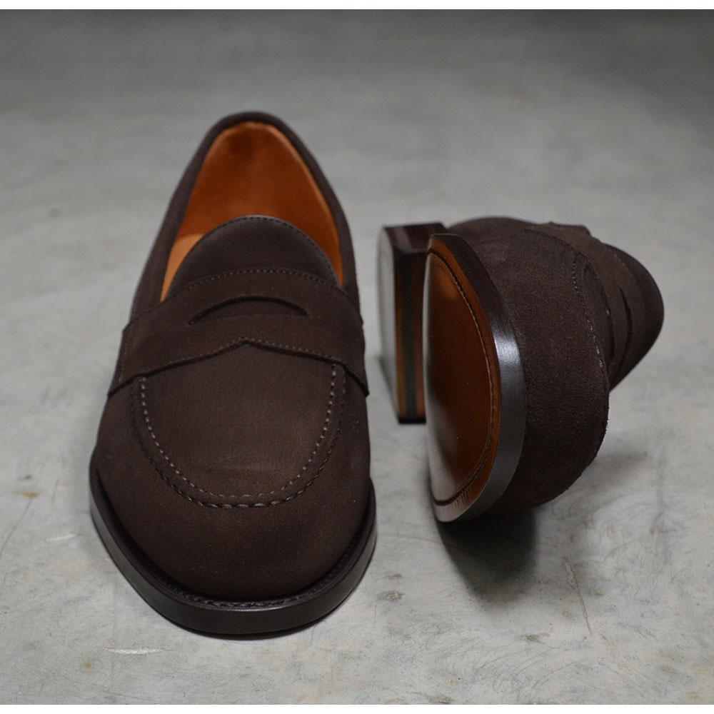 A dark brown suede penny loafer with a Goodyear welted leather sole, displayed on a polished concrete background.