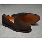 A dark brown suede penny loafer with a Goodyear welted leather sole, displayed on a polished concrete background.