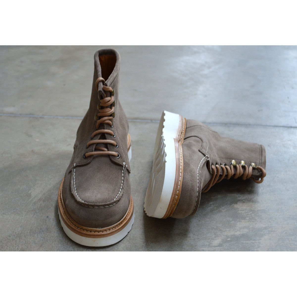 A grey and tan suede apron boot with lace-up closure, featuring a white sole and a natural welt.