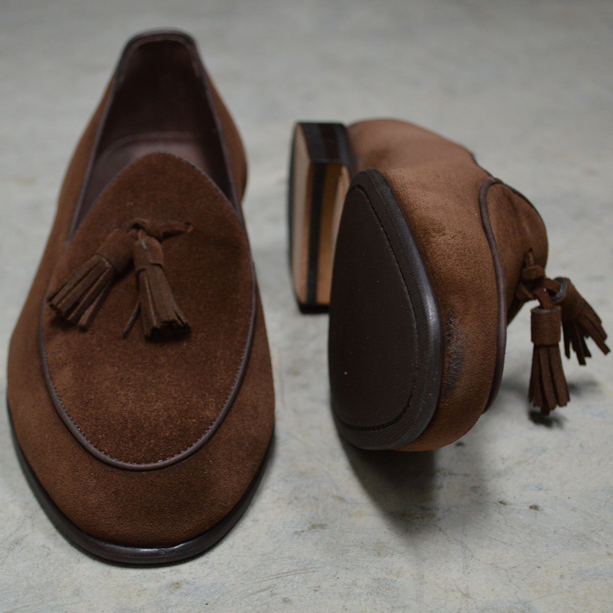 A dark brown suede tassel loafer with a Vegano Moka leather trim, displaying an almond-shaped toe and a fine ridged rubber sole.